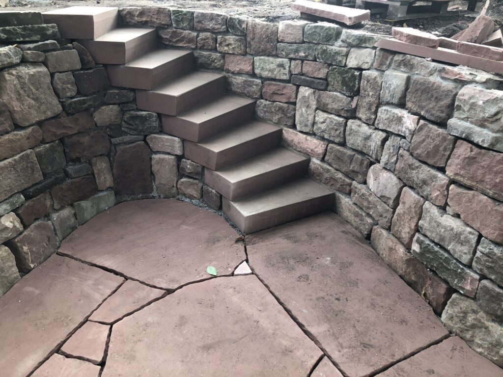 Sie sehen eine gerundete Natursteinmauer aus Sandstein mit einer gewendelten Treppe in die Mauer eingelassen.